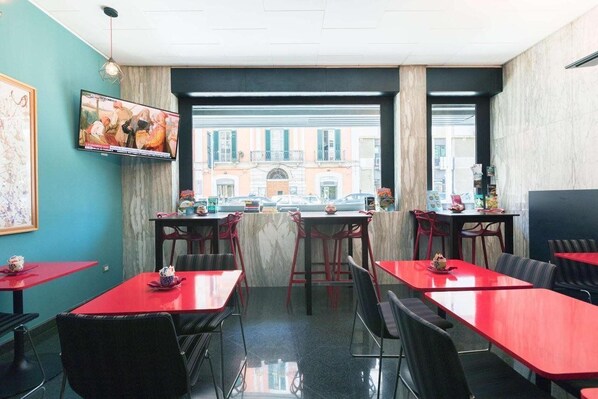 A restaurant with red tables and black chairs, a TV mounted on the wall, and a view of buildings outside.