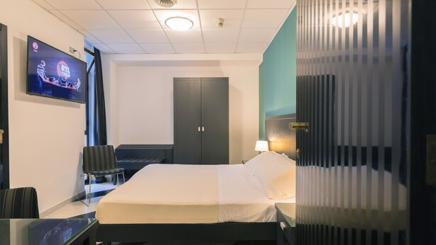 Hotellihuone, jossa on sänky, työpöytä, tuoli, televisio, vaatekaappi ja ikkuna, jossa on sälekaihtimet.