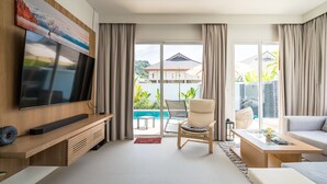 A modern living room with a flat-screen TV, wooden entertainment unit, and a large window overlooking a pool and greenery. - Kamala Garden View villa by Lofty  (Kamala)