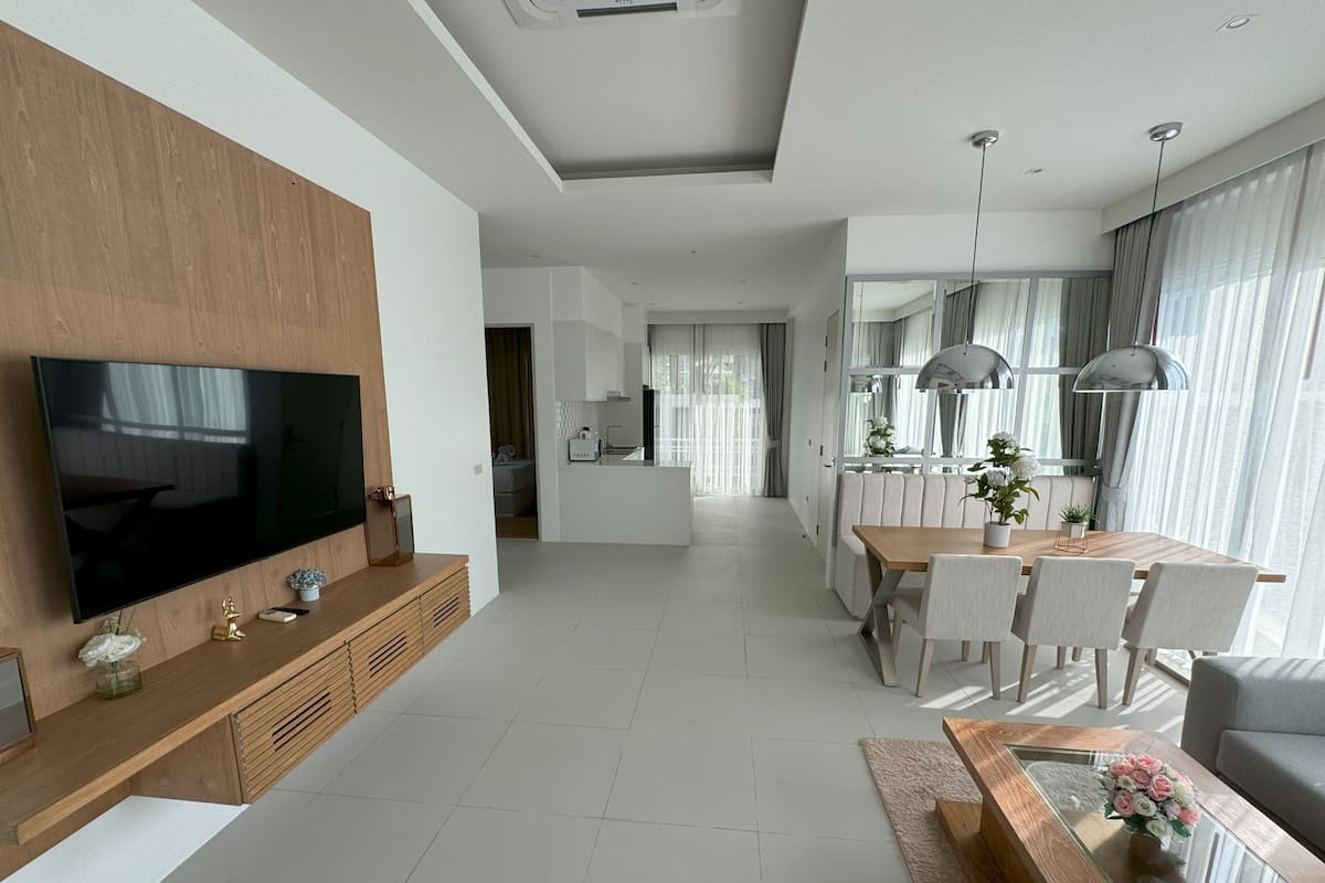 Una sala de estar moderna con televisor de pantalla plana, una consola de madera, comedor con mesa de vidrio y una cocina al fondo.