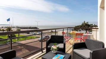 A balcony with wicker furniture, a table with a book, and a view of the sea and parking area.