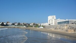 Une zone côtière avec une plage de sable, un grand bâtiment blanc à plusieurs étages et un ciel bleu dégagé.