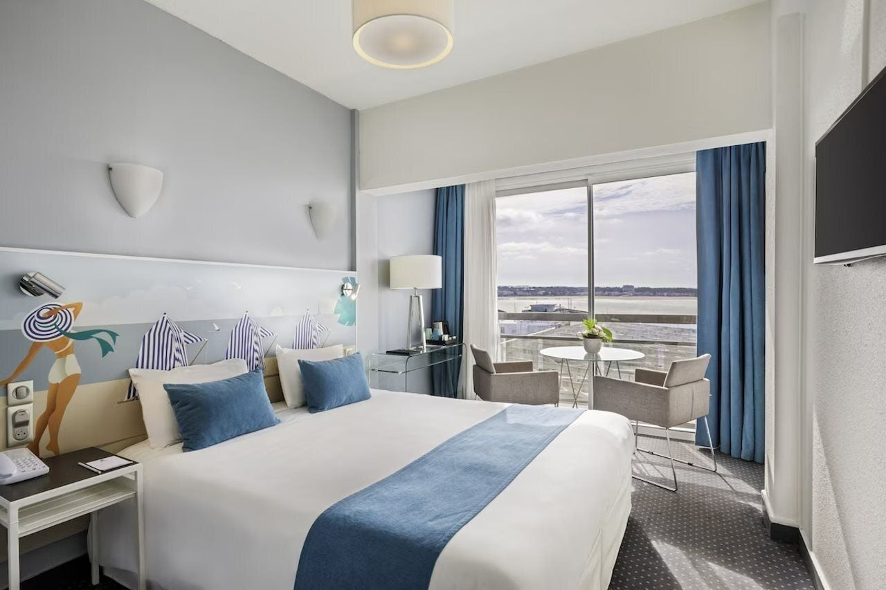 Quarto de hotel com uma cama grande, uma escrivaninha com abajur, uma pequena mesa com cadeiras e vista para a praia.