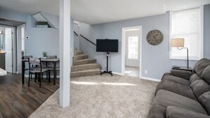 A living room with a gray couch, a TV on a stand, a staircase, and a dining area with a table and chairs. - This property is conviently located in Lakeside, within minutes from the Lake. (Muskegon)