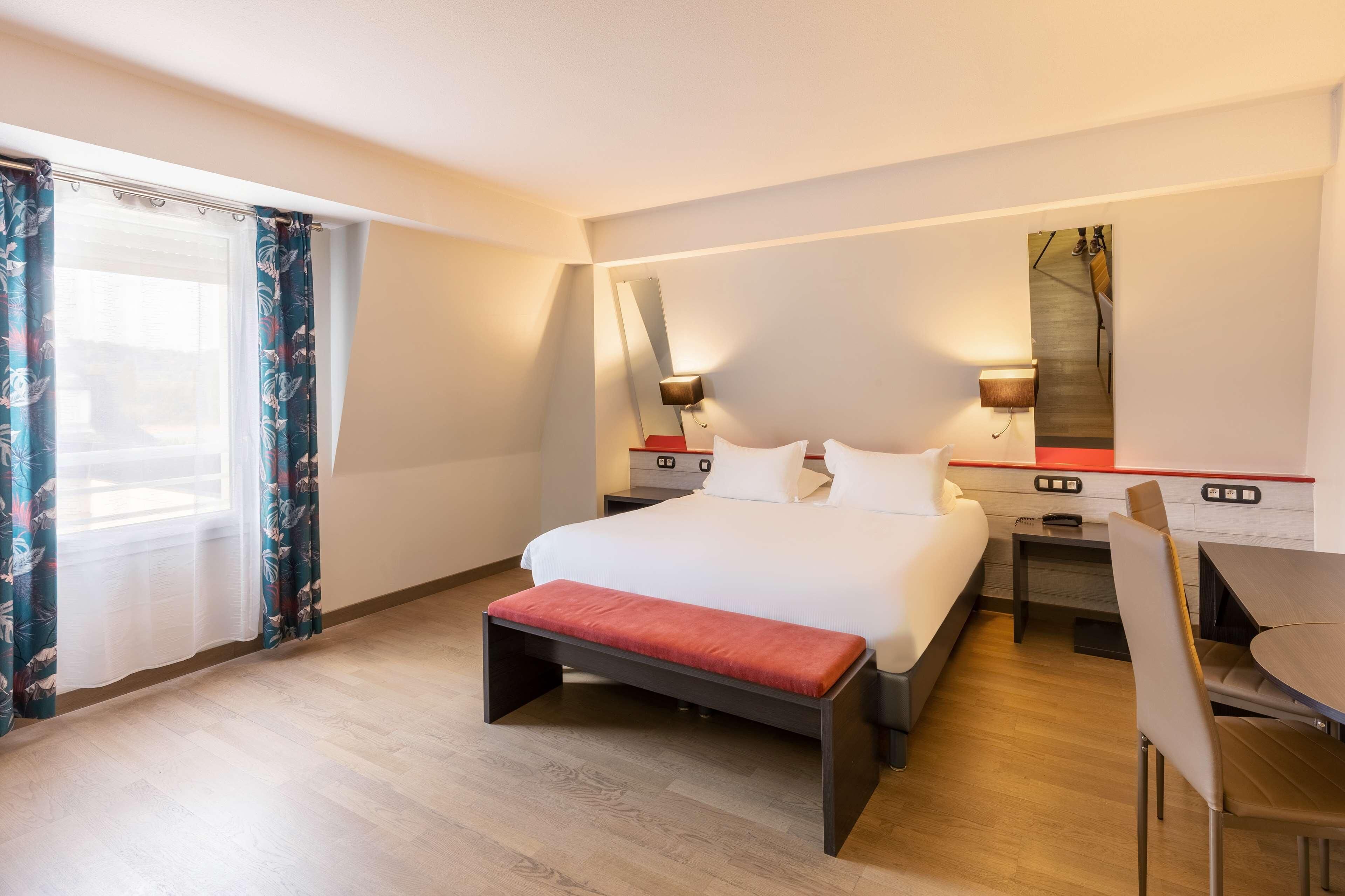 A hotel room with a bed, a desk, a chair, a bench, a nightstand, and a wall-mounted lamp.