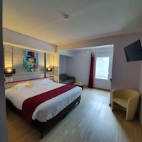 Quarto de hotel com uma cama grande, uma escrivaninha, uma cadeira, um quadro na parede e uma janela com cortinas.