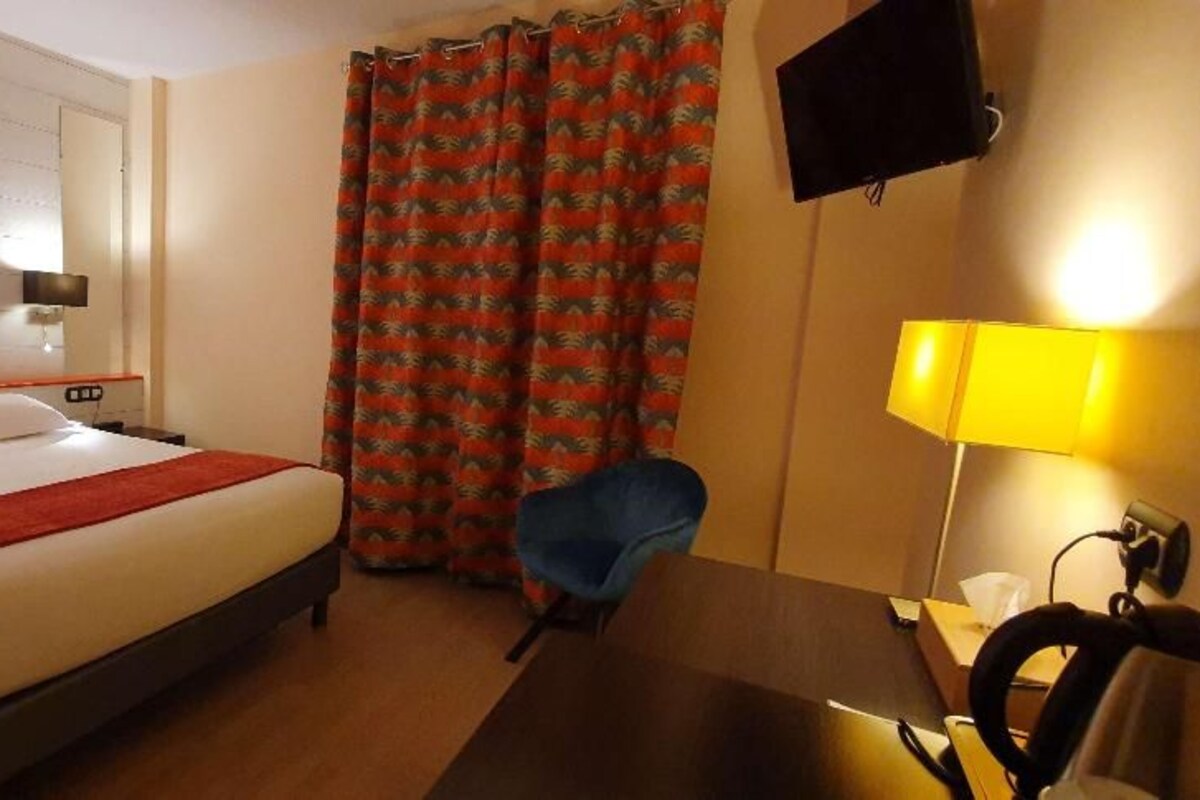 Habitación de hotel con cama, escritorio con lámpara, silla, televisor montado en la pared y ventana con cortinas.