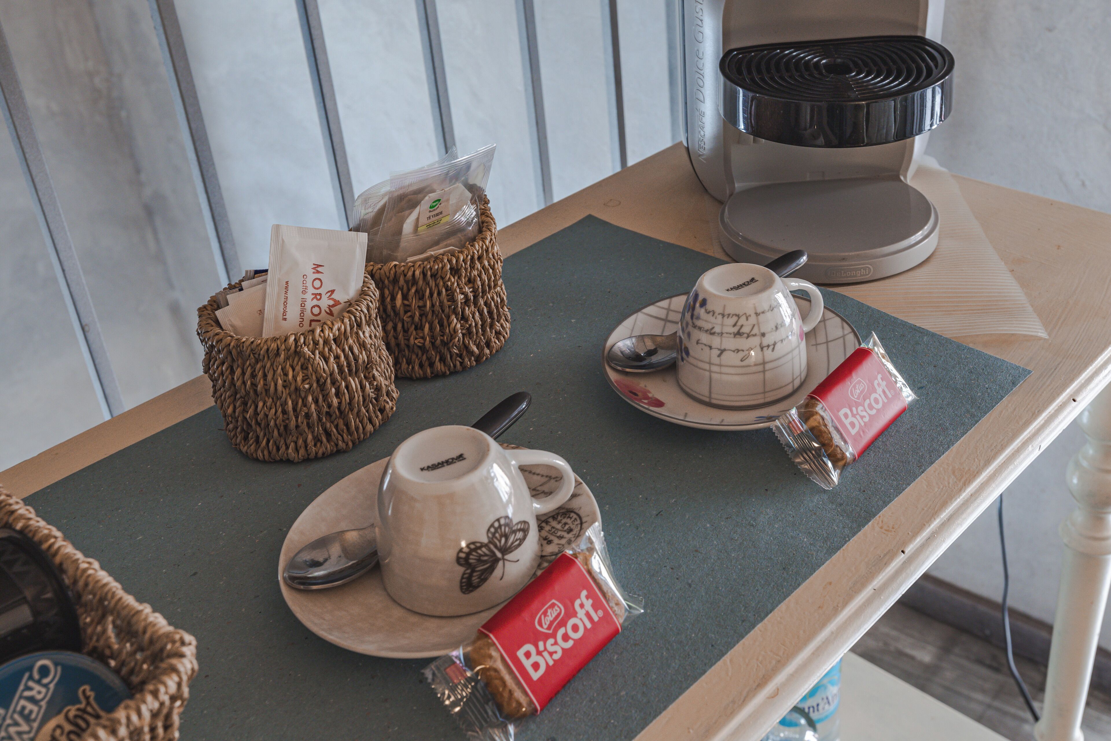 Una mesa con una cafetera, una taza con cuchara, un plato con una galleta y una cesta con café envasado.