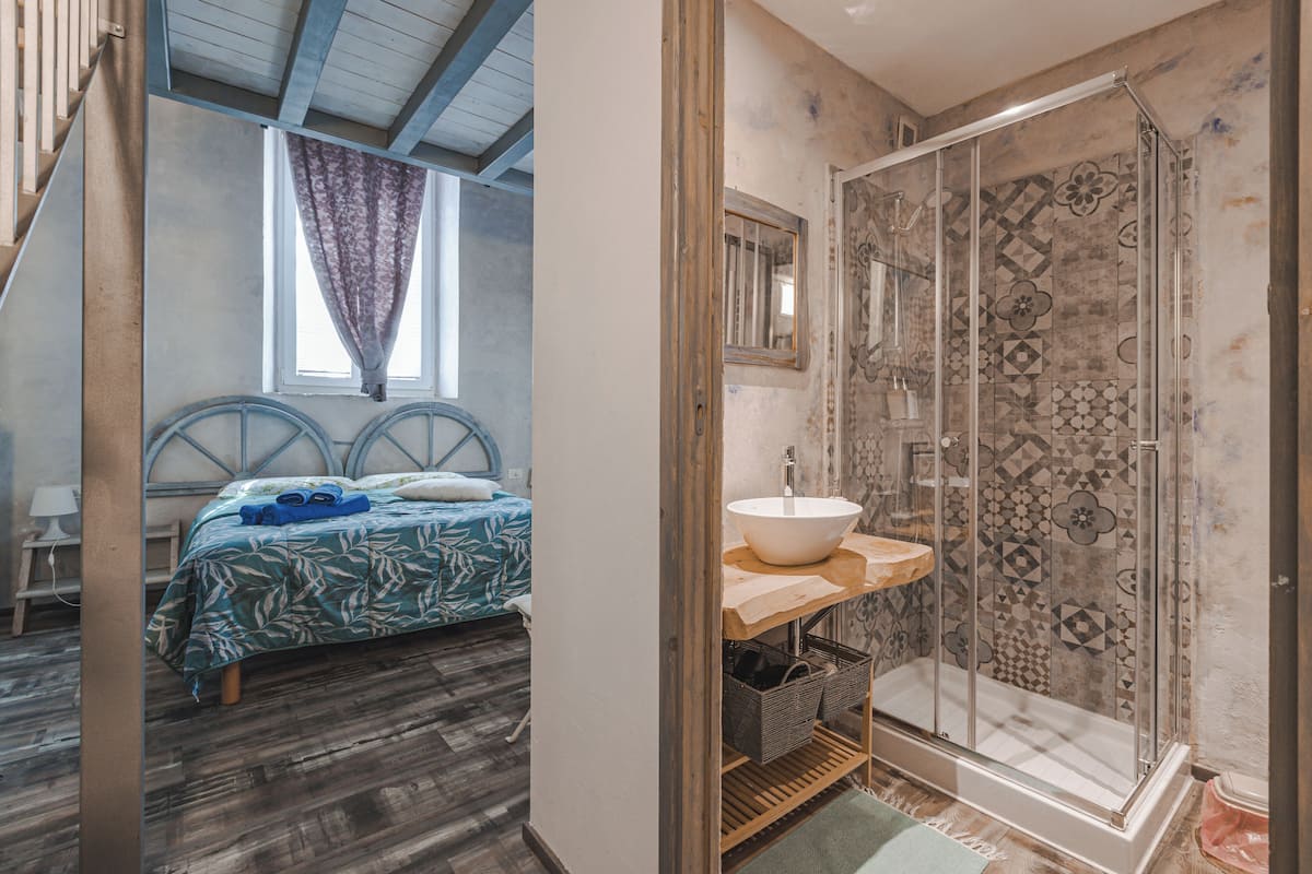 A bedroom with a bed, a window with curtains, and a bathroom with a sink and shower.
