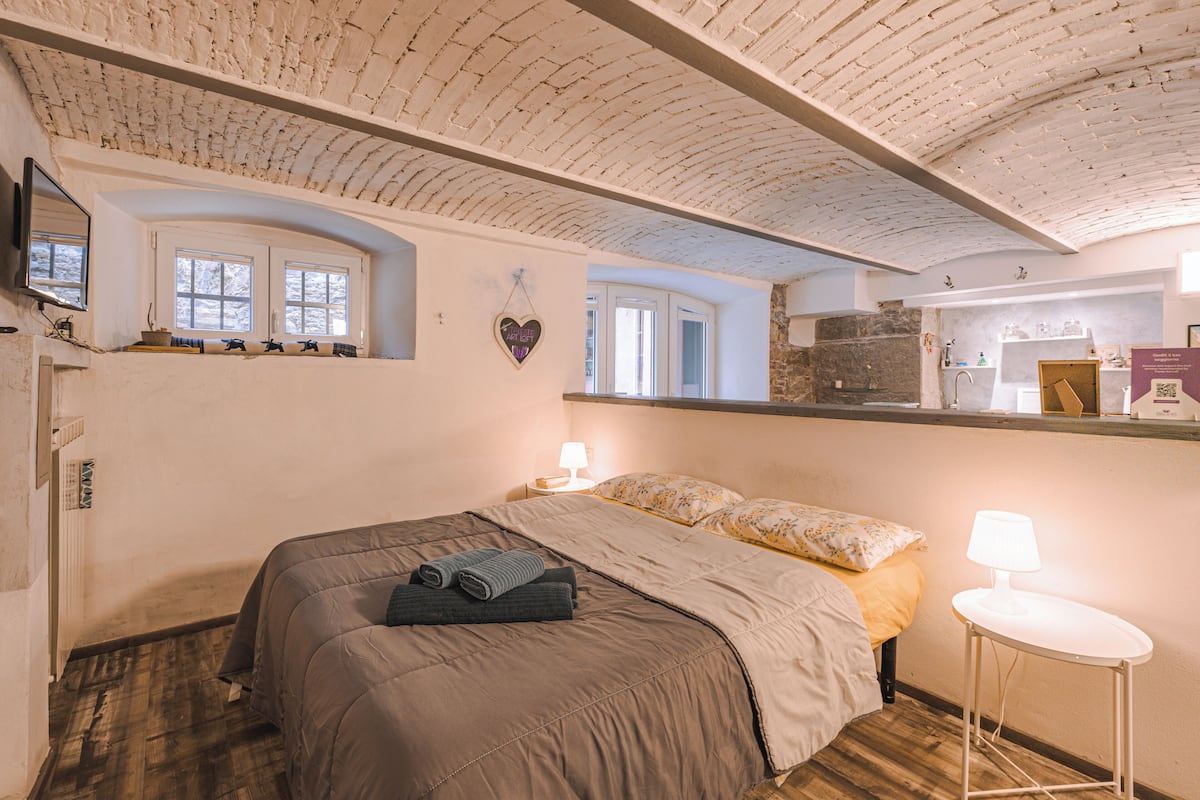 A bedroom with a large bed, a bedside table with a lamp, a heart-shaped decoration on the wall, and a window with a view of the outside.