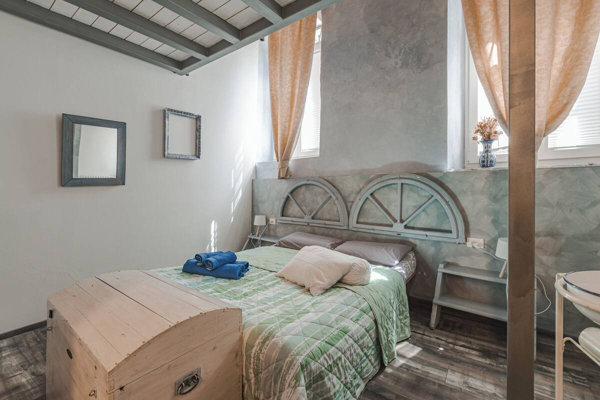 A bedroom with a wooden chest, a bed with a green quilt, two bedside tables, a nightstand, and a window with curtains.