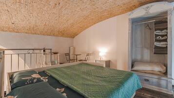 A bedroom with a wooden ceiling, a bed with floral bedding, a bedside table with a lamp, and a wardrobe with hanging clothes.