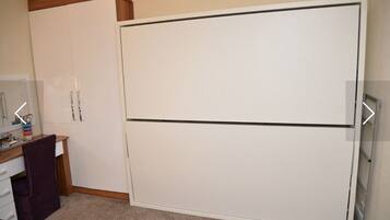 A room with a white wardrobe, a desk with a chair, and a mirror.
