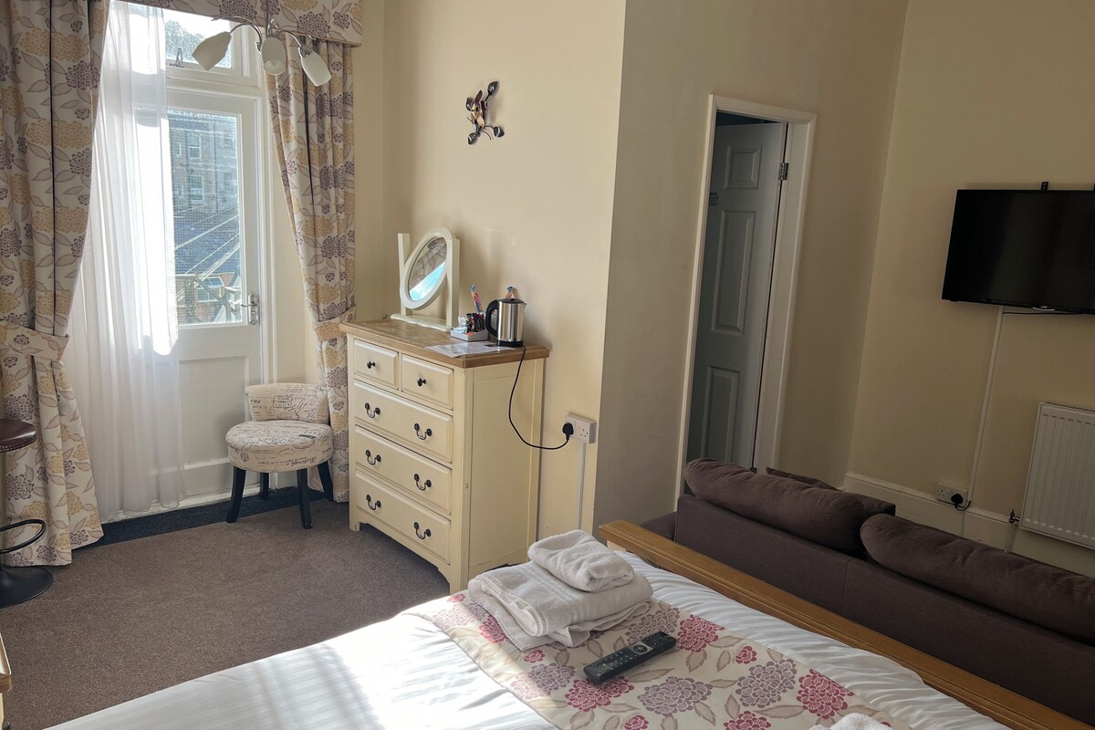 A bedroom with a bed, a dresser, a chair, a sofa, a TV, and a window with curtains.