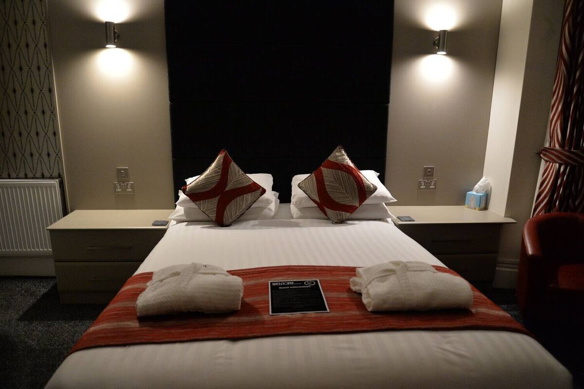 A hotel room with a bed, two bedside tables, a nightstand, a red and white striped blanket, two towels, a welcome card, and two wall-mounted lights.