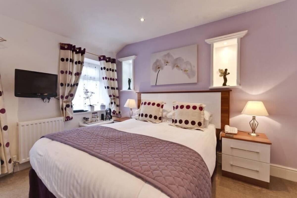 A bedroom with a large bed, a bedside table with a lamp, a TV mounted on the wall, and a window with curtains.