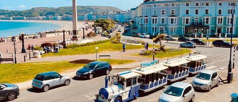 A coastal street with parked cars, a tram, and a prominent monument.