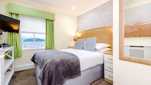 A bedroom with a bed, a TV, a window with curtains, a headboard, and a wall with a geometric pattern. - Lawton & Lauriston Court (Llandudno)