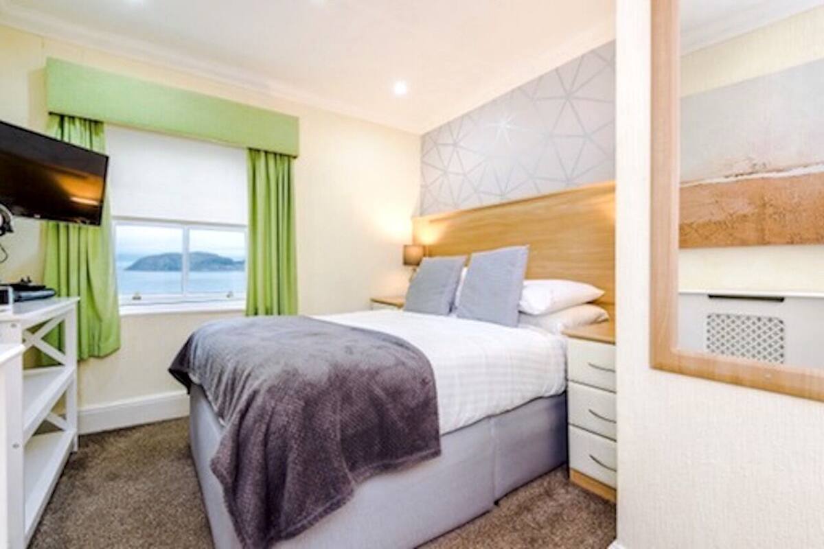 A bedroom with a bed, a TV, a window with curtains, a headboard, and a wall with a geometric pattern.