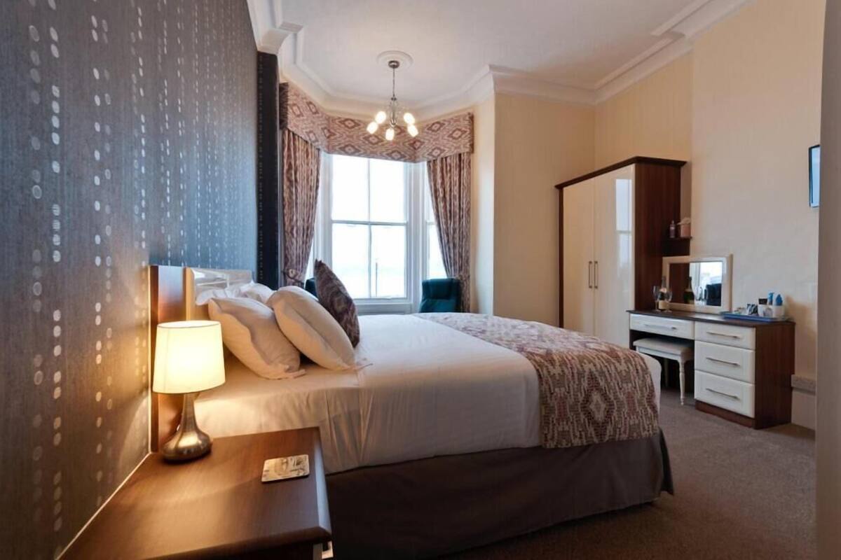 A hotel room with a large bed, a desk, a chair, a lamp, and a wardrobe.