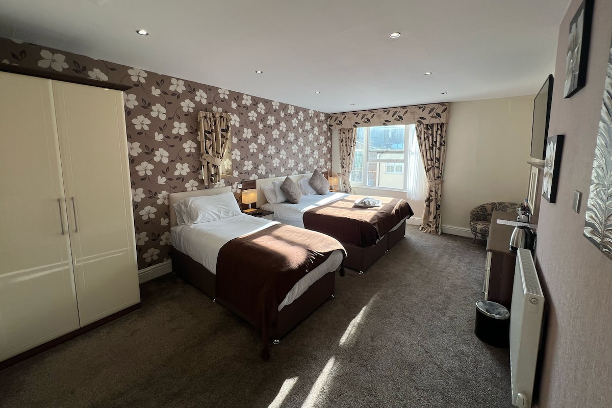A hotel room with two beds, a wardrobe, a radiator, and a window with curtains.