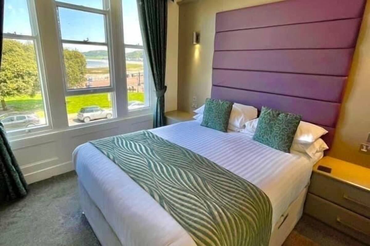A hotel room with a large bed, a view of the outdoors through a window, and a purple headboard.