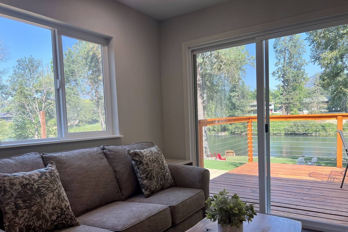 A modern living room with a grey sofa, patterned cushions, and a wooden coffee table with a plant. Large windows provide a view of a deck and outdoor area.