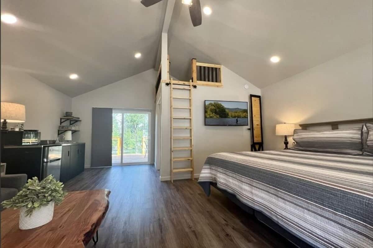 A bedroom with a lofted sleeping area, a wooden ladder, a bed with striped bedding, a desk with a lamp, and a view of greenery through a door.