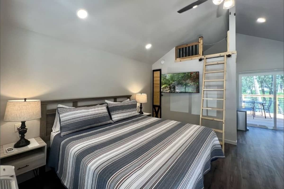 A bedroom with a large bed, a nightstand, a desk, a ladder, and a view of the outdoors.