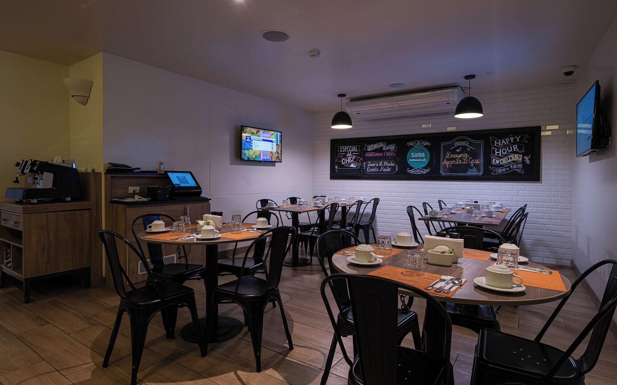 Interior de restaurante con varias mesas y sillas, una máquina de café, una pantalla de televisión y un menú en pizarra.