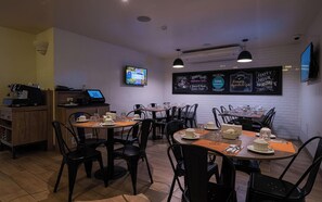 Interior de restaurante con varias mesas y sillas, una máquina de café, una pantalla de televisión y un menú en pizarra.