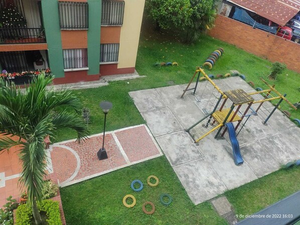 Une aire de jeux avec un toboggan et une structure d’escalade, entourée d’une pelouse verdoyante et d’un mur de briques.