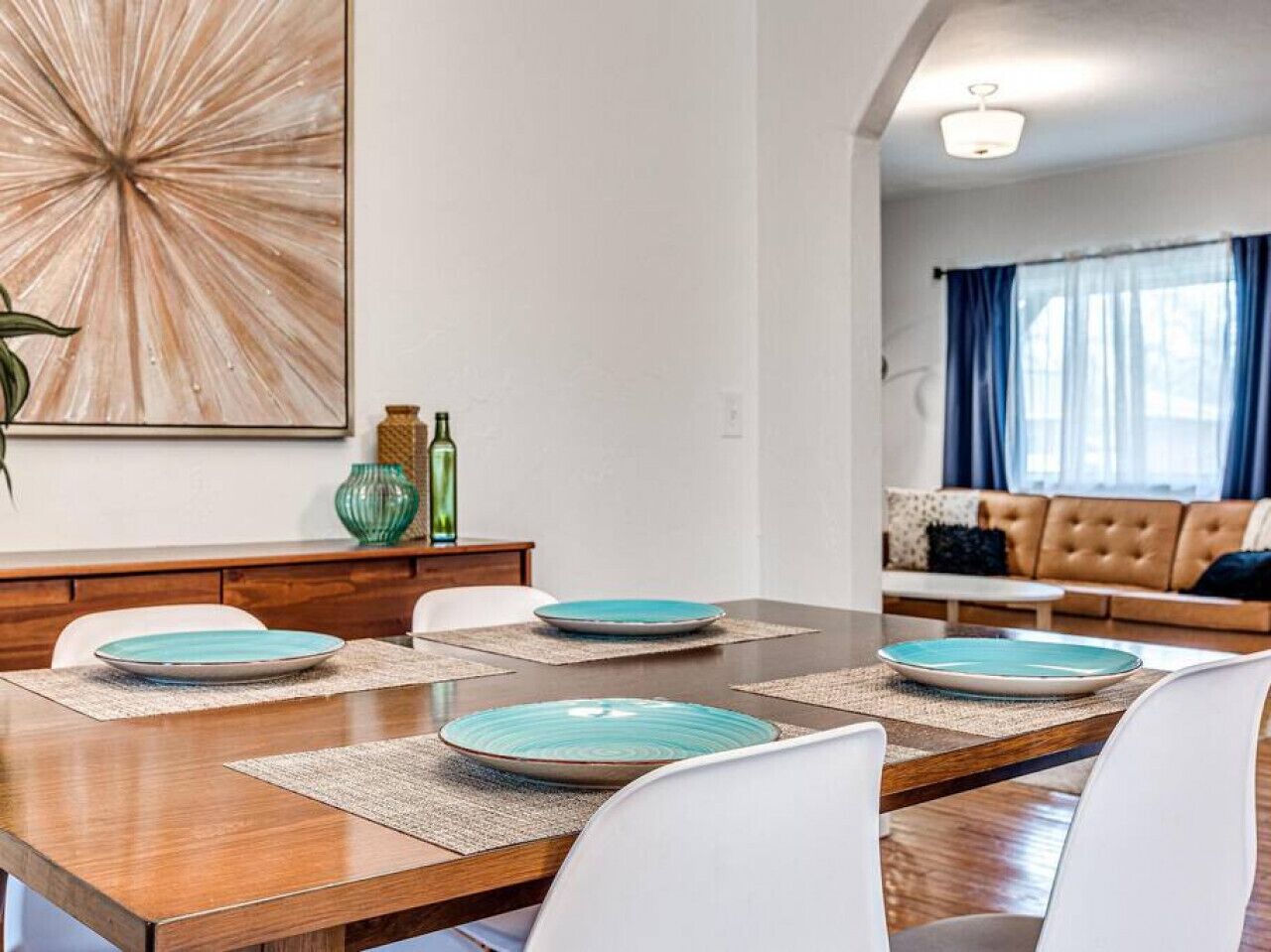 A dining area with a wooden table, white chairs, and blue plates. There is a sofa and a window with blue curtains in the background.