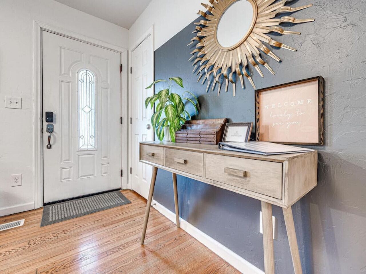 Een hal met een houten vloer, een witte deur met een ruit, een console tafel met lades, een potplant, een decoratieve spiegel en een welkomstbord.