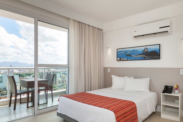 A hotel room with a bed, a desk, a chair, a balcony with a view of the city, and an air conditioning unit. - Ramada by Wyndham Macae Hotel & Suites (Macaé)