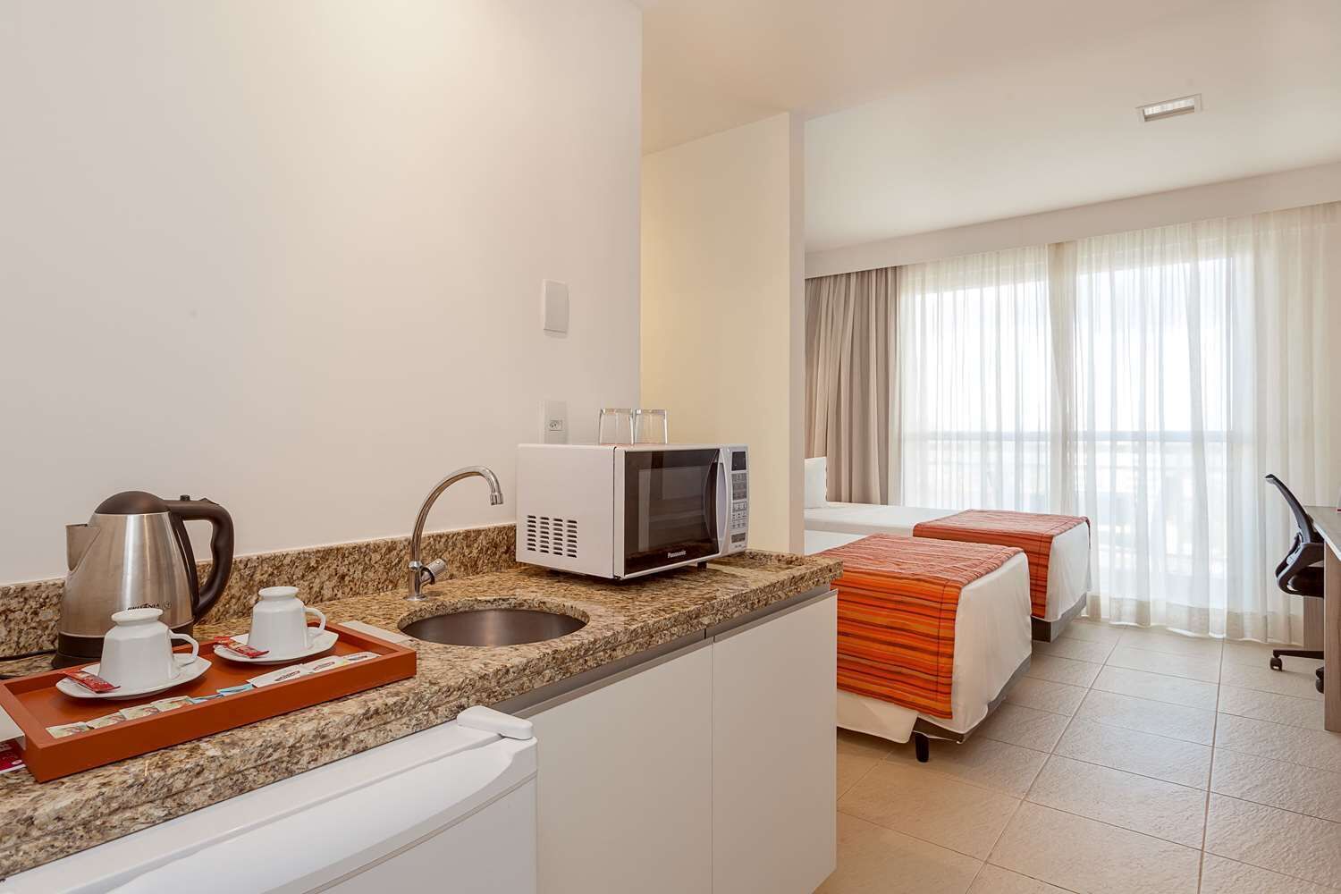 A hotel room with a kitchenette, a bed, a desk, and a view of the city.