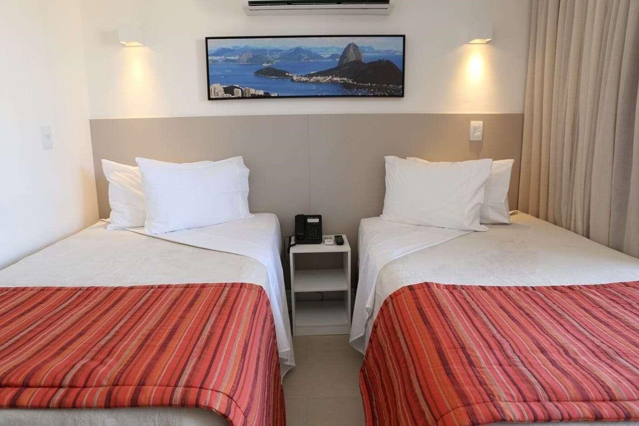 A hotel room with two beds, each with white bedding and red-striped bedspreads. There is a small bedside table with a phone and a lamp. A large framed picture of a coastal landscape hangs above the beds.