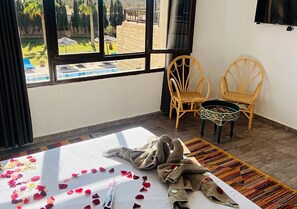 Ein Zimmer mit einem Bett, zwei Korbstühlen, einem Fernseher und Blick auf einen Pool und Grünflächen.