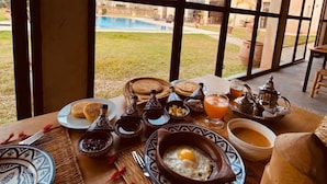 A breakfast table set with traditional Moroccan dishes, including a tagine, mint tea, and bread, in a room with large windows overlooking a pool. - Guesthouse Dar Ayman Essaouira B&B (Essaouira)