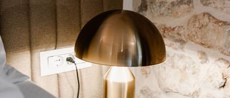 A lamp with a metallic shade on a bedside table, a power outlet on the wall, and a stone wall in the background.