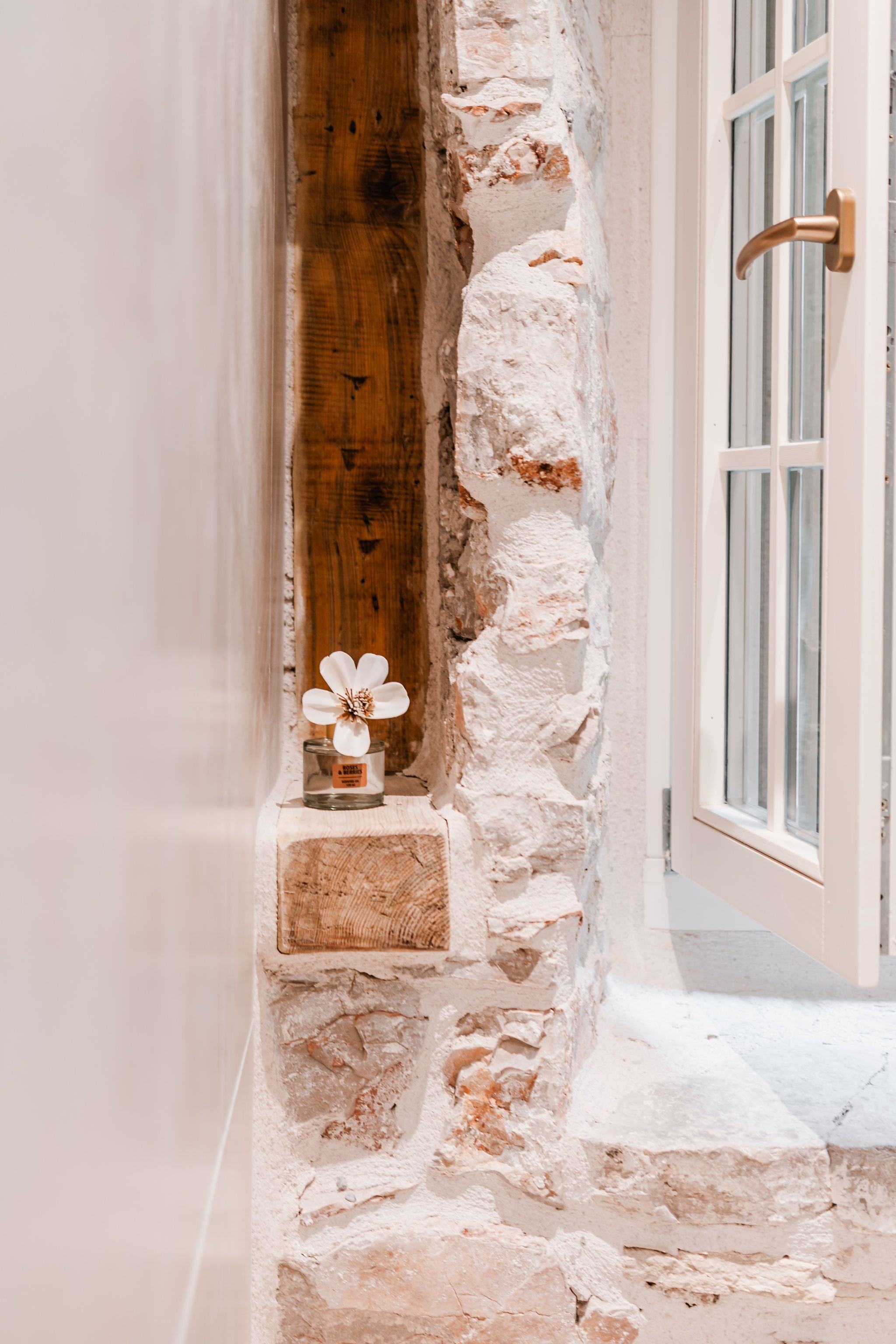 Un mur de pierres avec une poutre en bois, une porte blanche avec une poignée en laiton, et une petite table sur laquelle sont posées une fleur blanche et une bougie.