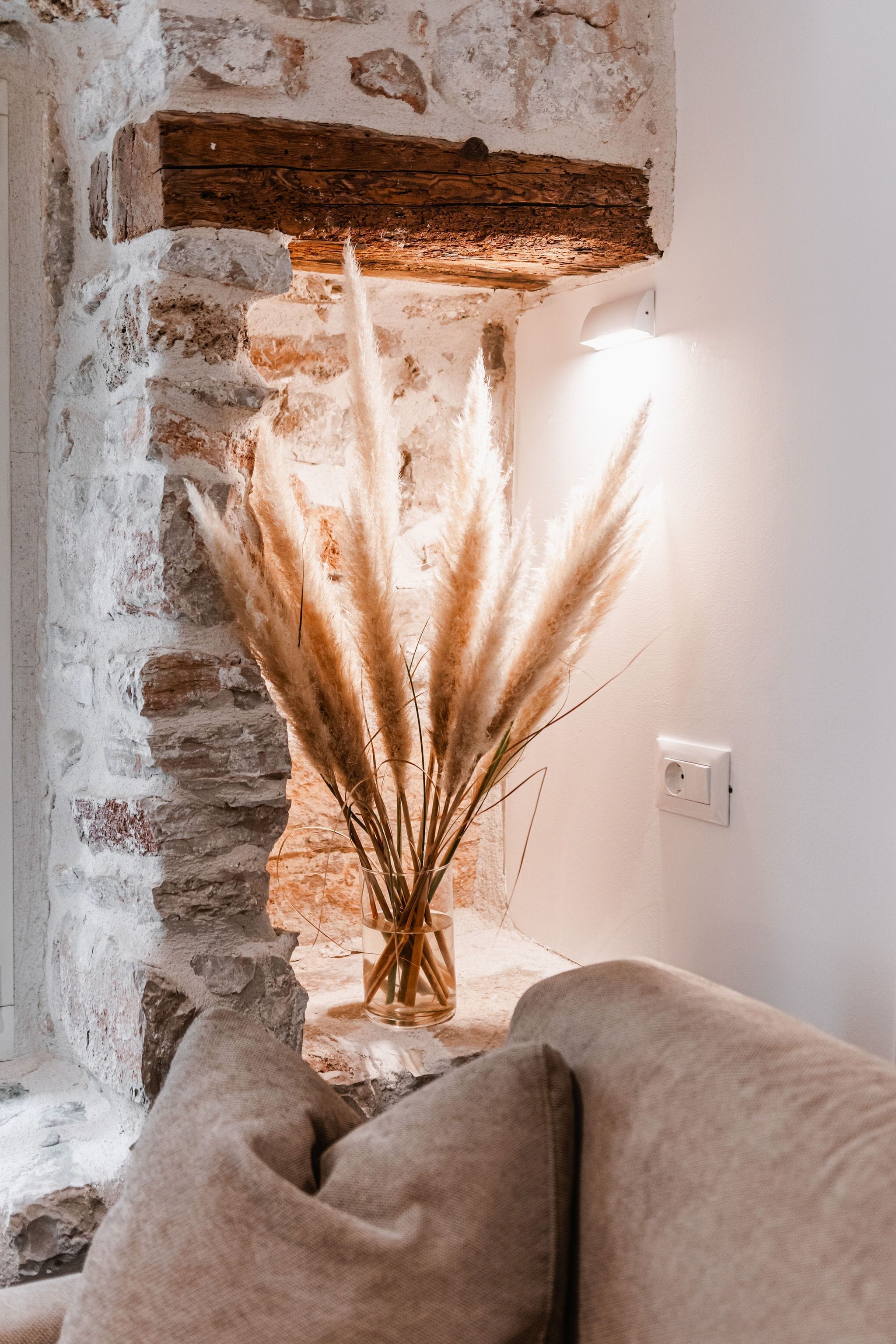 Un intérieur rustique avec un mur en pierre, une poutre en bois et un vase contenant des plumes d’herbe de la pampa.