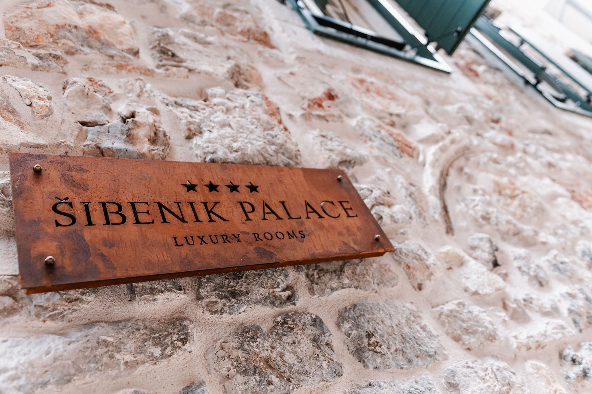 A wooden sign with "Sibenik Palace Luxury Rooms" written on it, mounted on a stone wall.