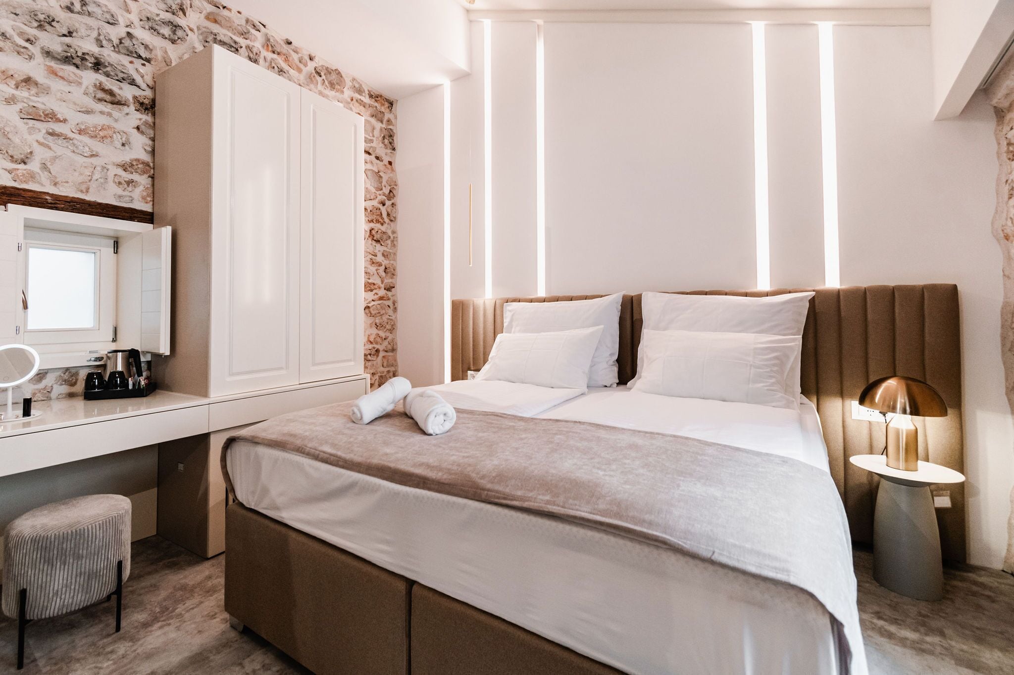 A hotel room with a stone wall, a large bed with white bedding, a bedside table with a lamp, and a desk with a mirror and a coffee maker.