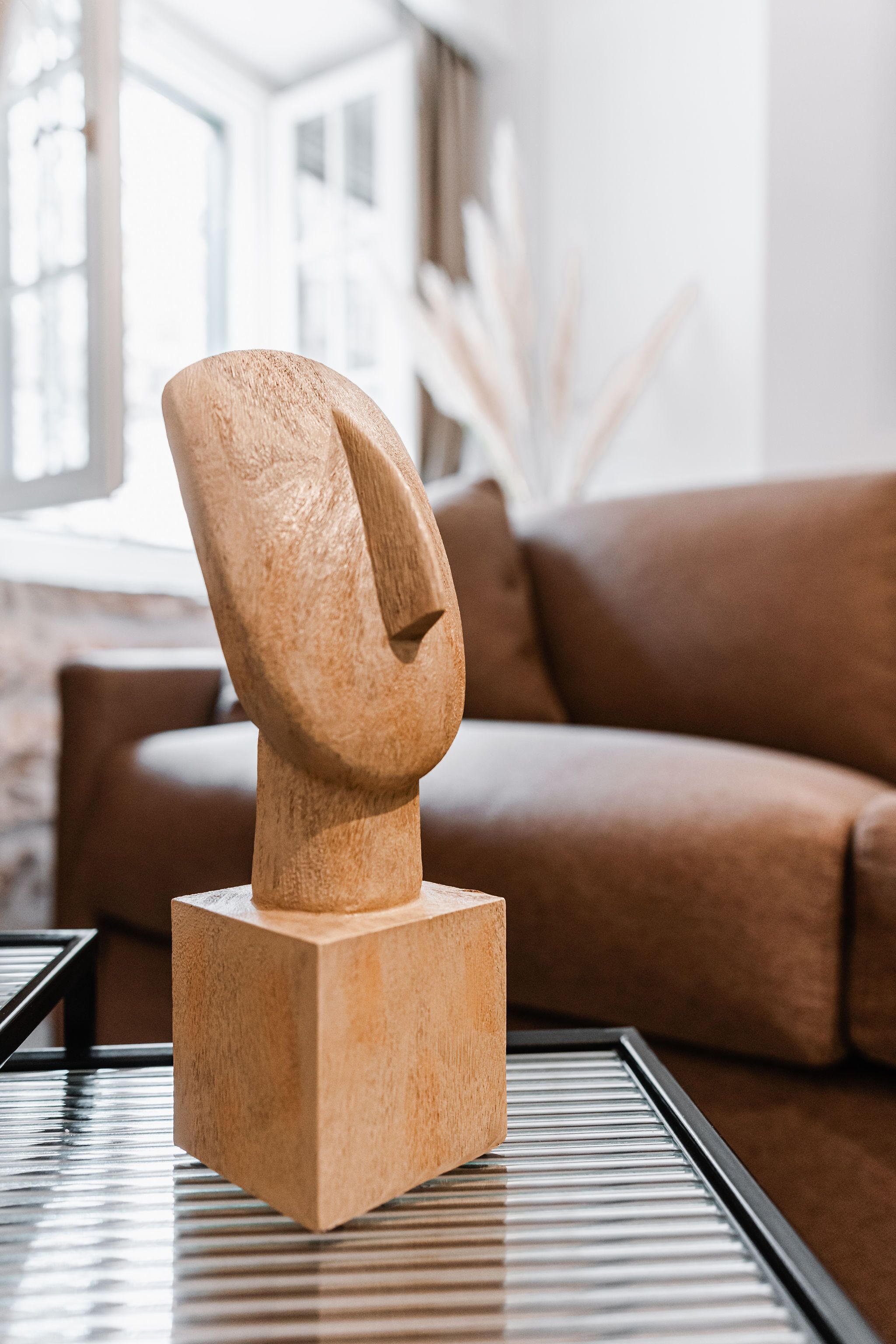 En moderne skulptur på et glastilbud i en stue med en brun sofa og et stort vindue i baggrunden.