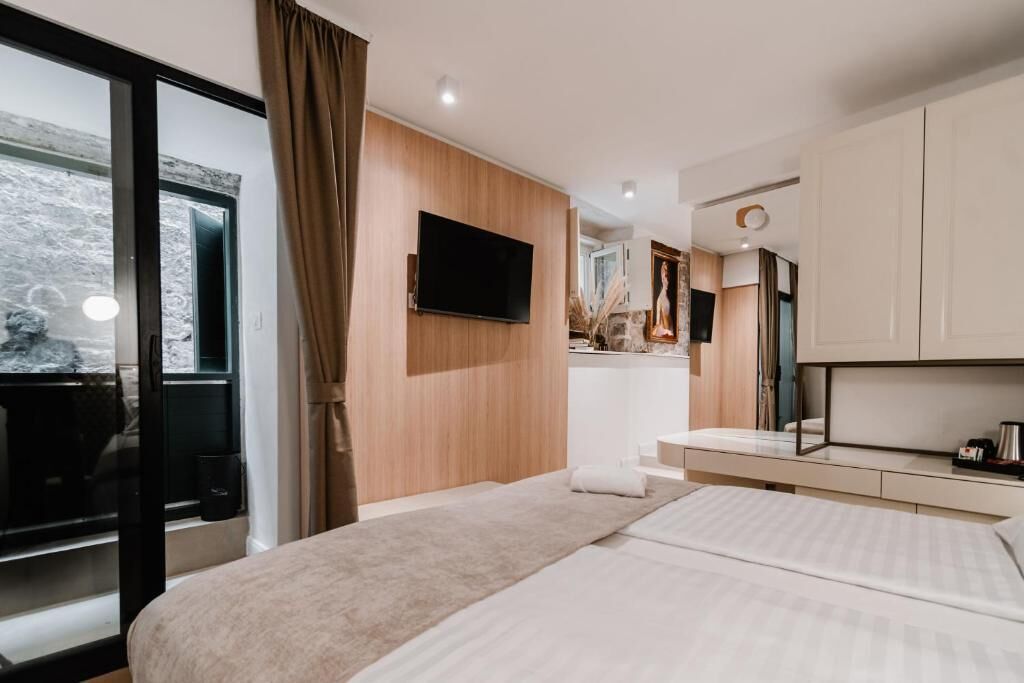 Una habitación de hotel moderna con una cama grande, un televisor de pantalla plana montado en la pared, una kitchenette pequeña y un ventanal corredizo que da a un balcón.