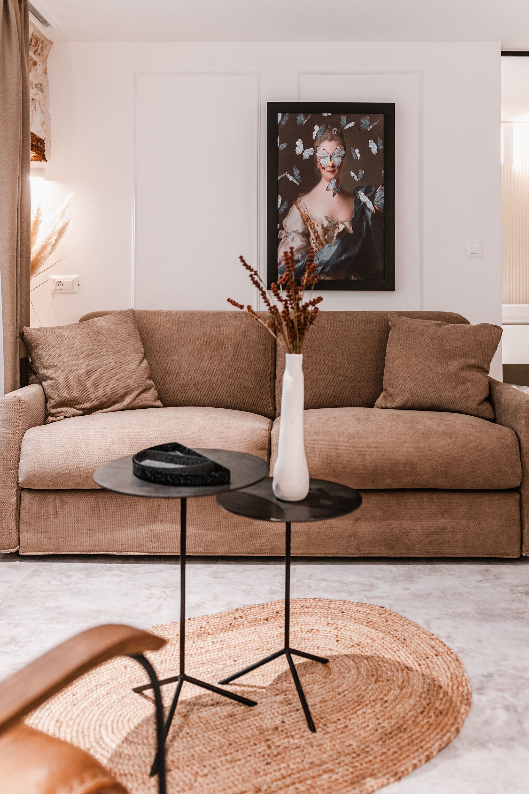 Una sala de estar moderna con un sofá color beige, un jarrón blanco con plantas secas y una obra de arte enmarcada en la pared.