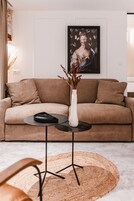 Una sala de estar moderna con un sofá color beige, un jarrón blanco con plantas secas y una obra de arte enmarcada en la pared.