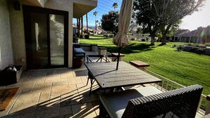 Un patio con tavolo e sedie, un ombrellone e vista su un prato curato e case sullo sfondo.