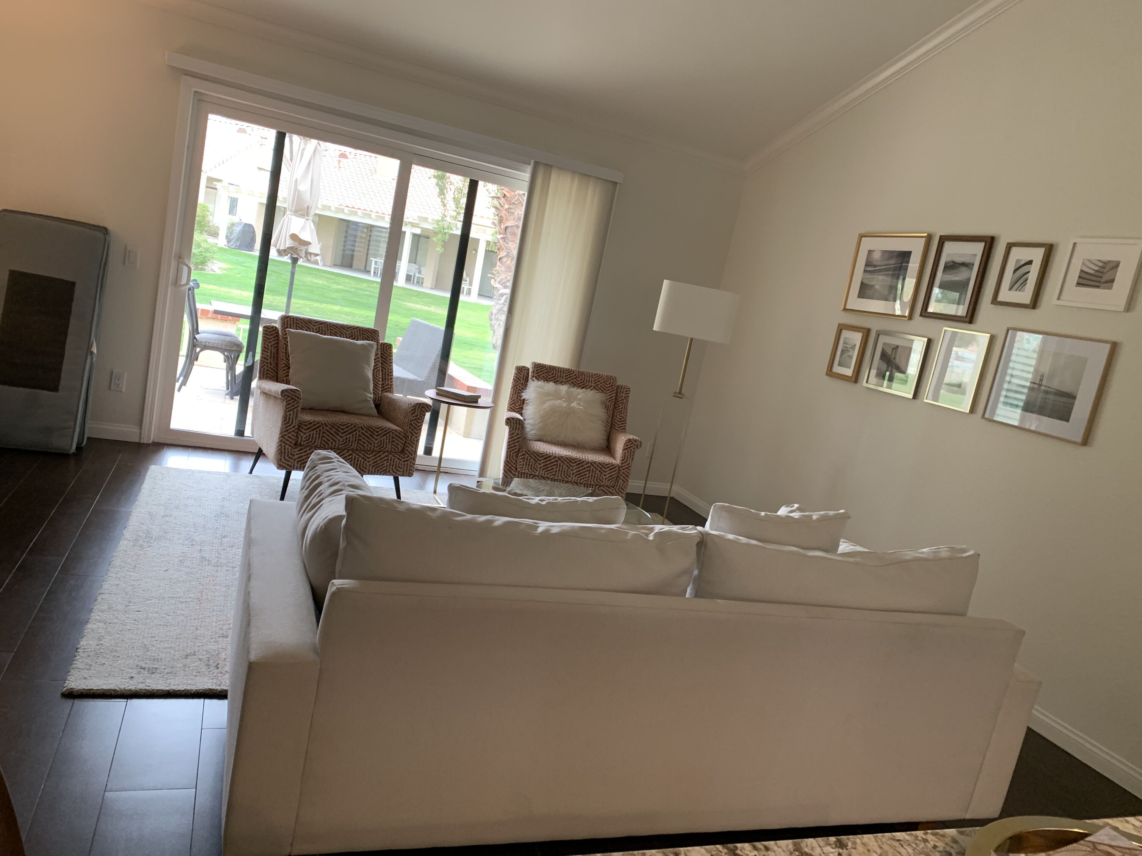A living room with a sofa, two patterned armchairs, a coffee table, and a TV. There are framed pictures on the wall and a sliding glass door leading to a patio.